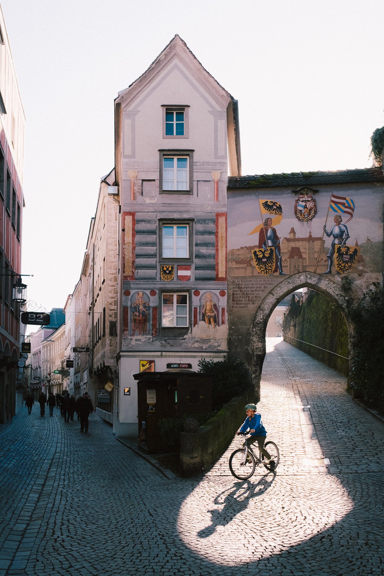 A cyclist rides through a painted medieval town gate, long shadows on the cobbles.