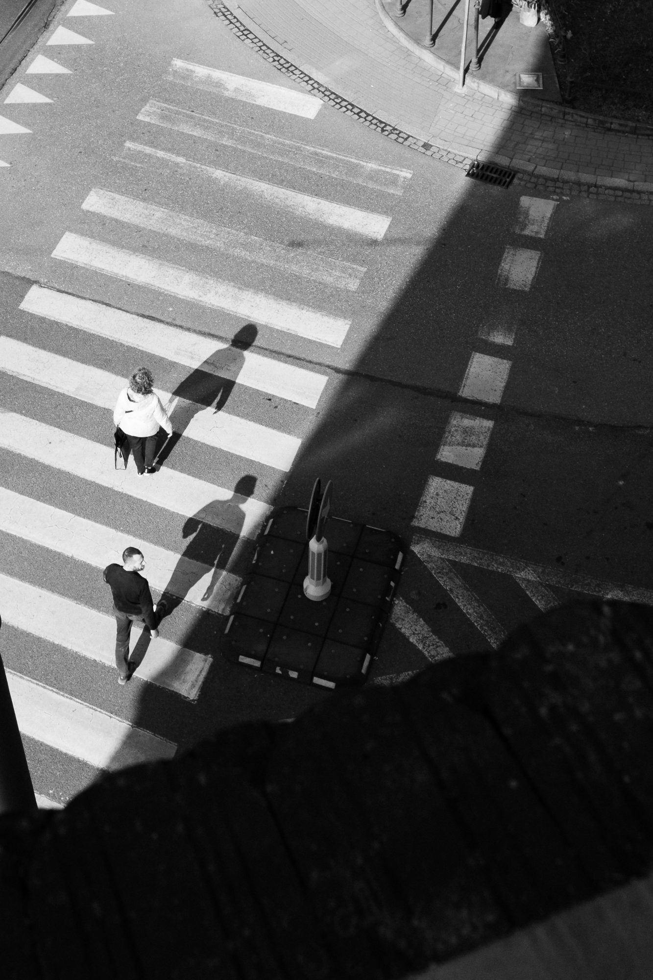 A zebra crossing from above, three people and their long shadows, a hard diagonal of shade.