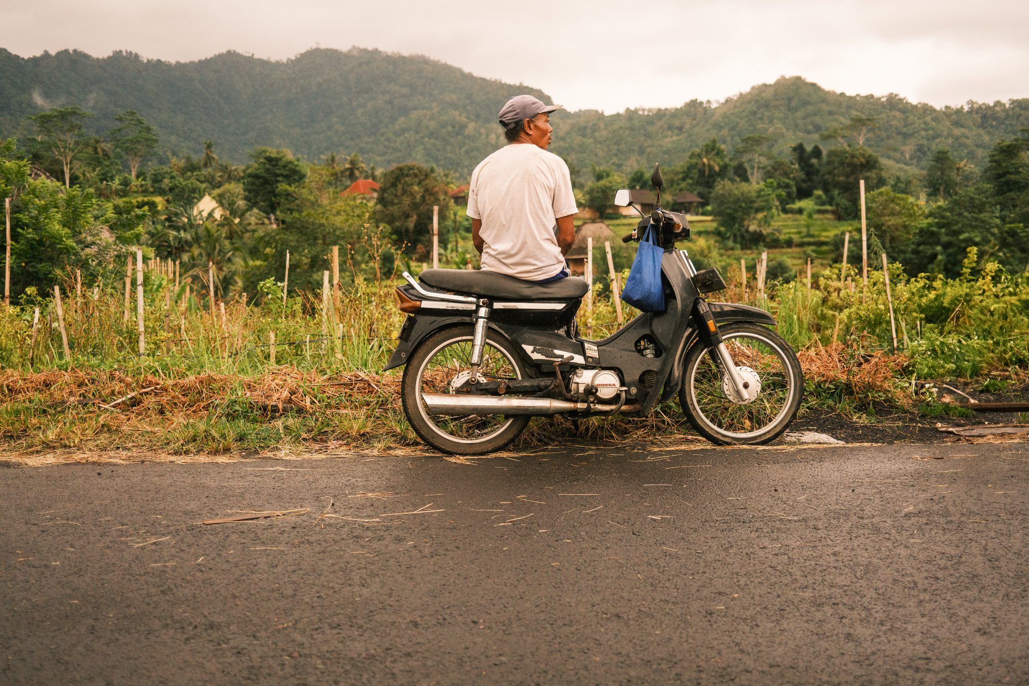 A man sits on a parked moped at the edge of a wet road, facing the fields.