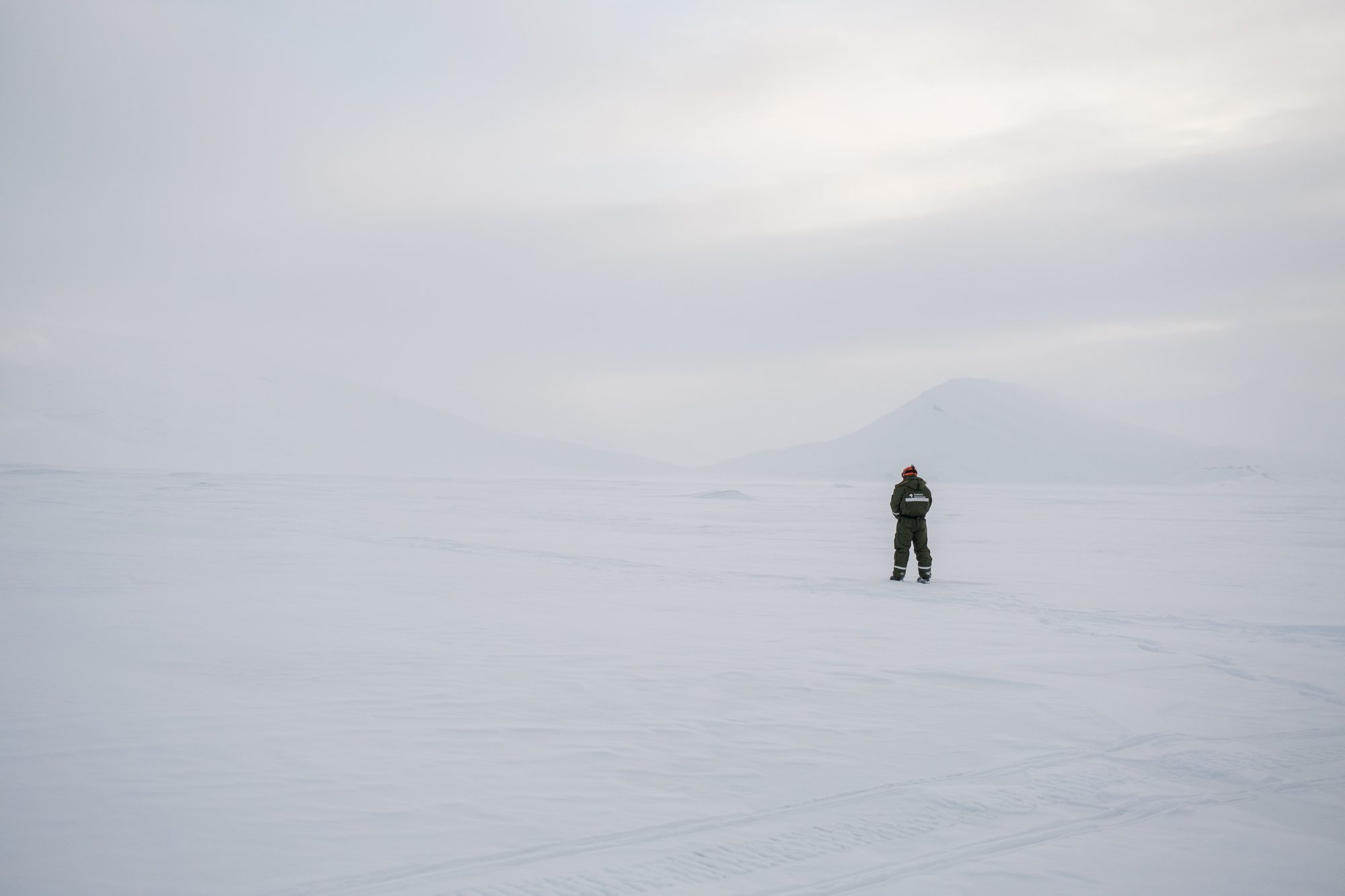 A lone figure stands small in a white snowfield under a pale sky.